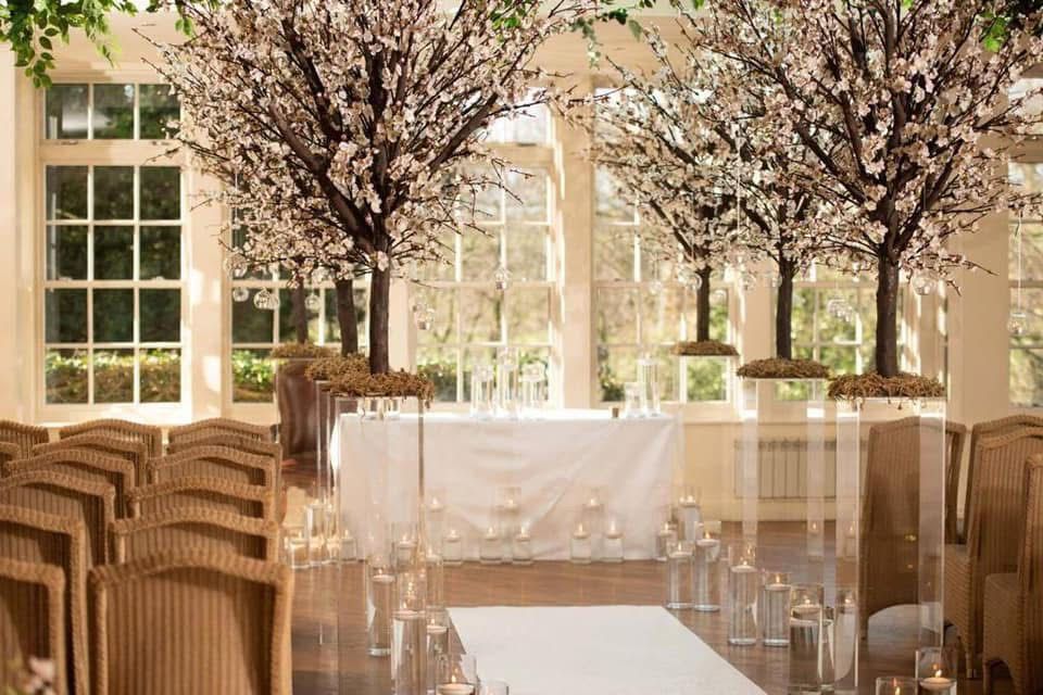 Wedding ceremony setup, light-filled room with blossoming trees as backdrop, white aisle runner, candles, and chairs.