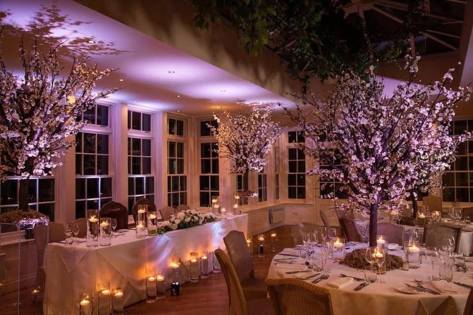 Formal dining room with white tablecloths, lit by candles and decorative blossoming trees.