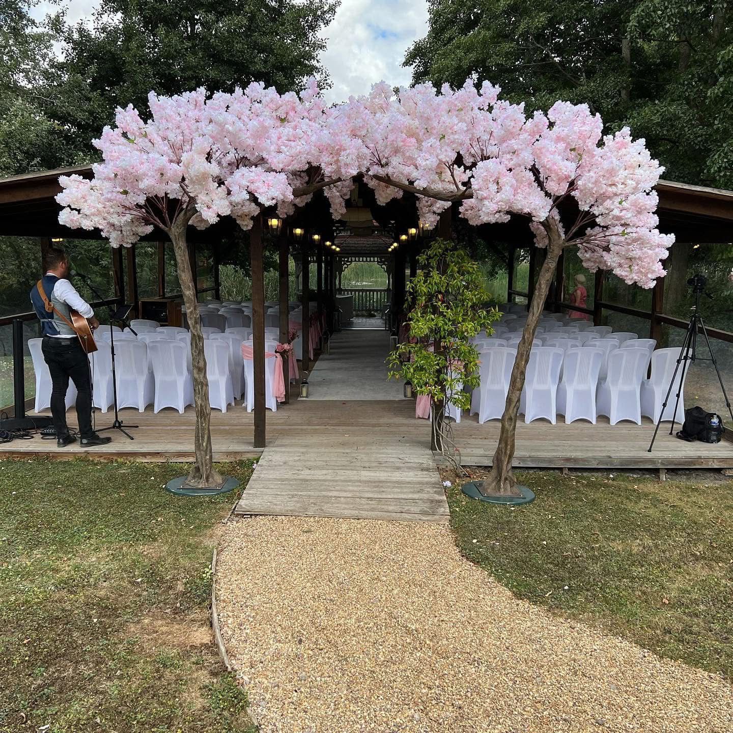 Wedding ceremony setting: cherry blossom trees frame aisle with rows of white chairs. Musician plays guitar.