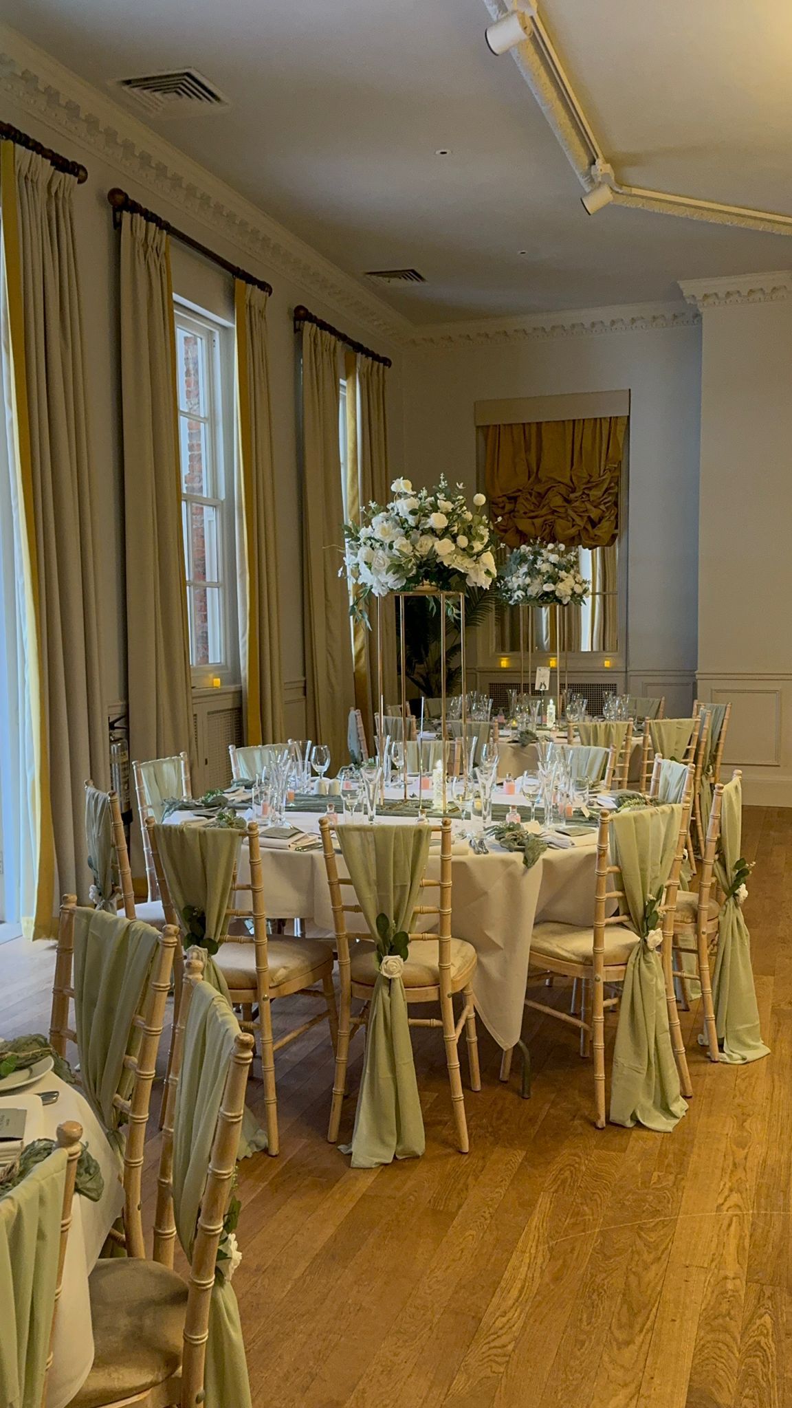 Formal dining table set for a wedding. Gold chairs, white linens, tall floral centerpieces, and draped green fabric.