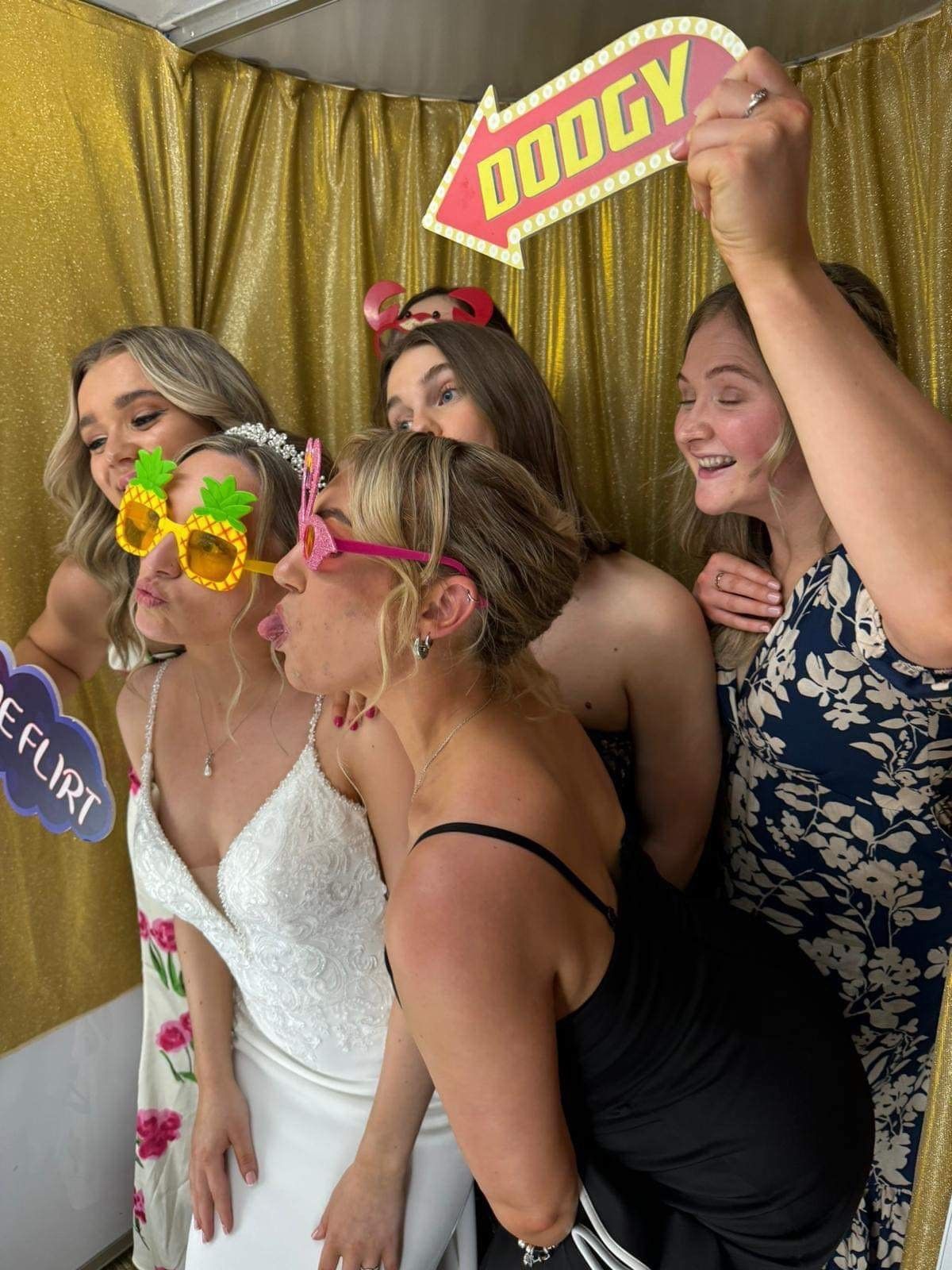 Six women pose in a photo booth with props. They are making kissing faces in front of a gold background.