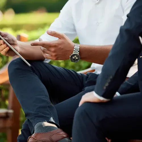 Two men in suits, one holding a tablet, gesturing outdoors.