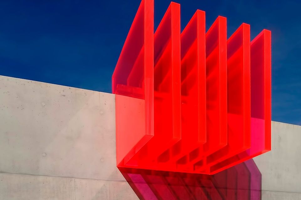 Bright red, geometric sculpture casting shadows on a concrete wall against a blue sky.