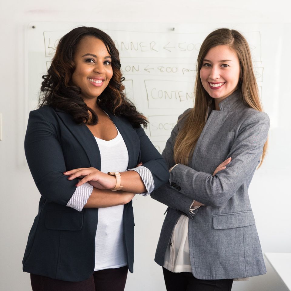 Two women, arms crossed, smiling. One is Black, wearing a blue blazer. The other is white, wearing a gray blazer.