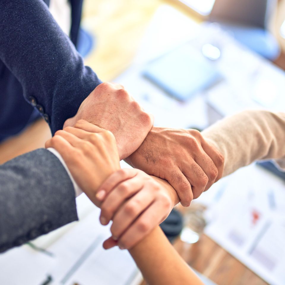 Four people joining hands in a team gesture, arms and hands in view, over a blurred office setting.