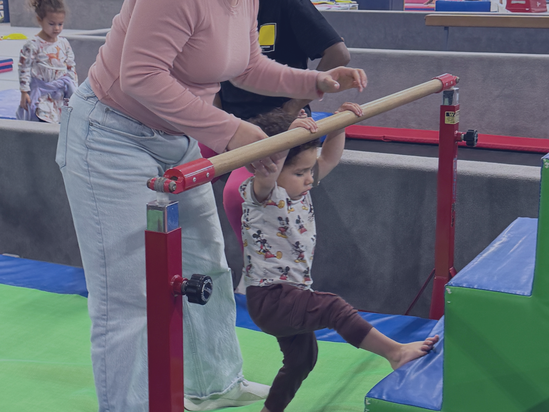 Toddler participating in a parent-tot gymnastics class, holding onto a low bar with assistance from 