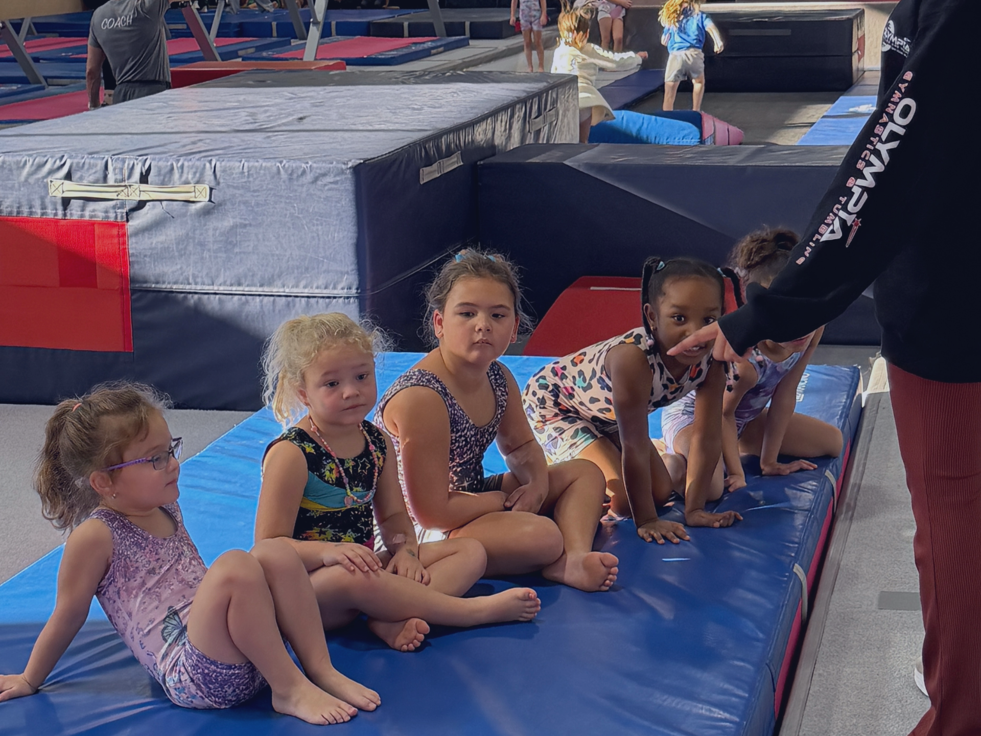 Gymnasts sitting on a mat inside a gymnastics facility while coach gives instructions.
