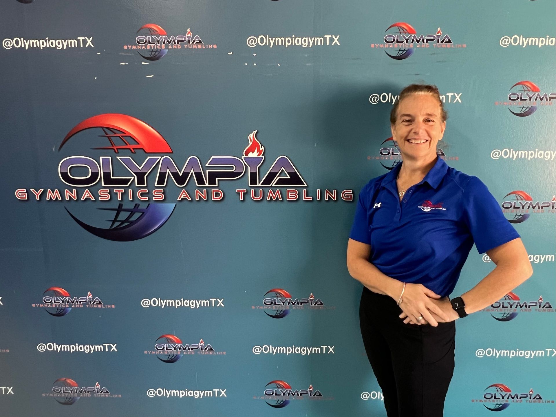 A woman in a blue shirt is standing in front of a wall with olympia gymnastics and tumbling written on it.