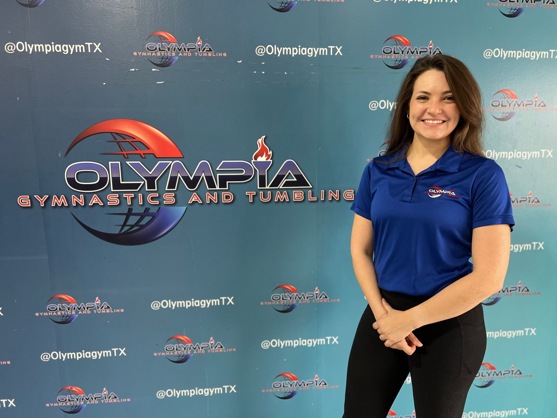 A woman in a blue shirt is standing in front of a wall that says olympia gymnastics and tumbling.