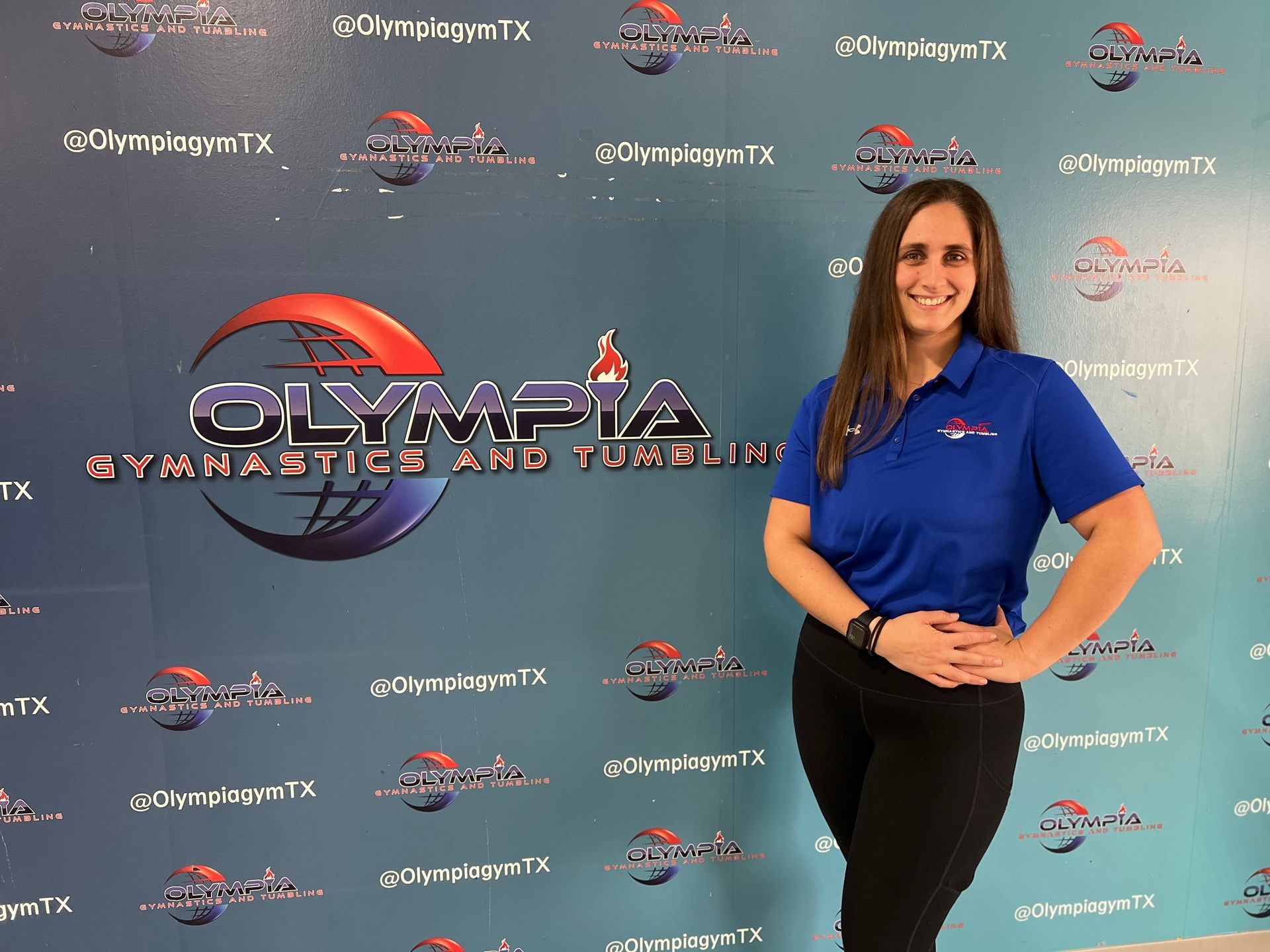 A woman in a blue shirt is standing in front of a wall that says olympia gymnastics and tumbling.
