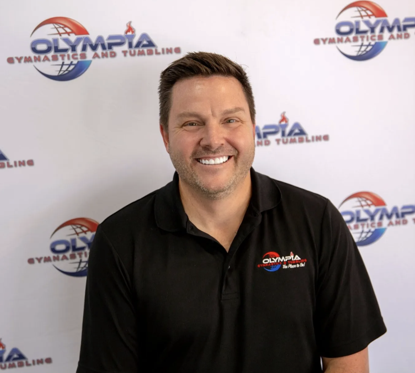 A man is smiling in front of a wall that says olympia gymnastics and tumbling