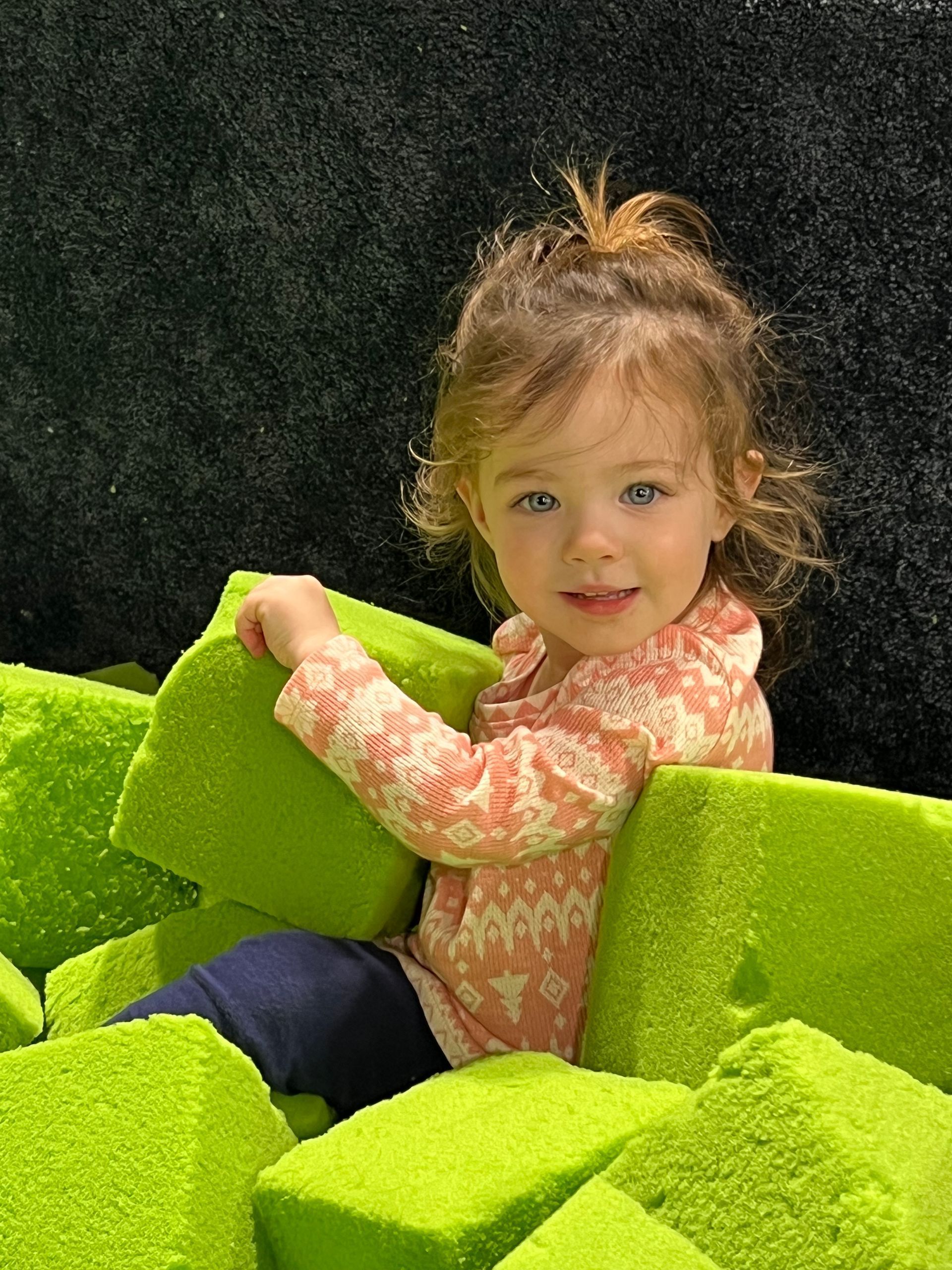 A little girl is sitting in a pile of green foam blocks.