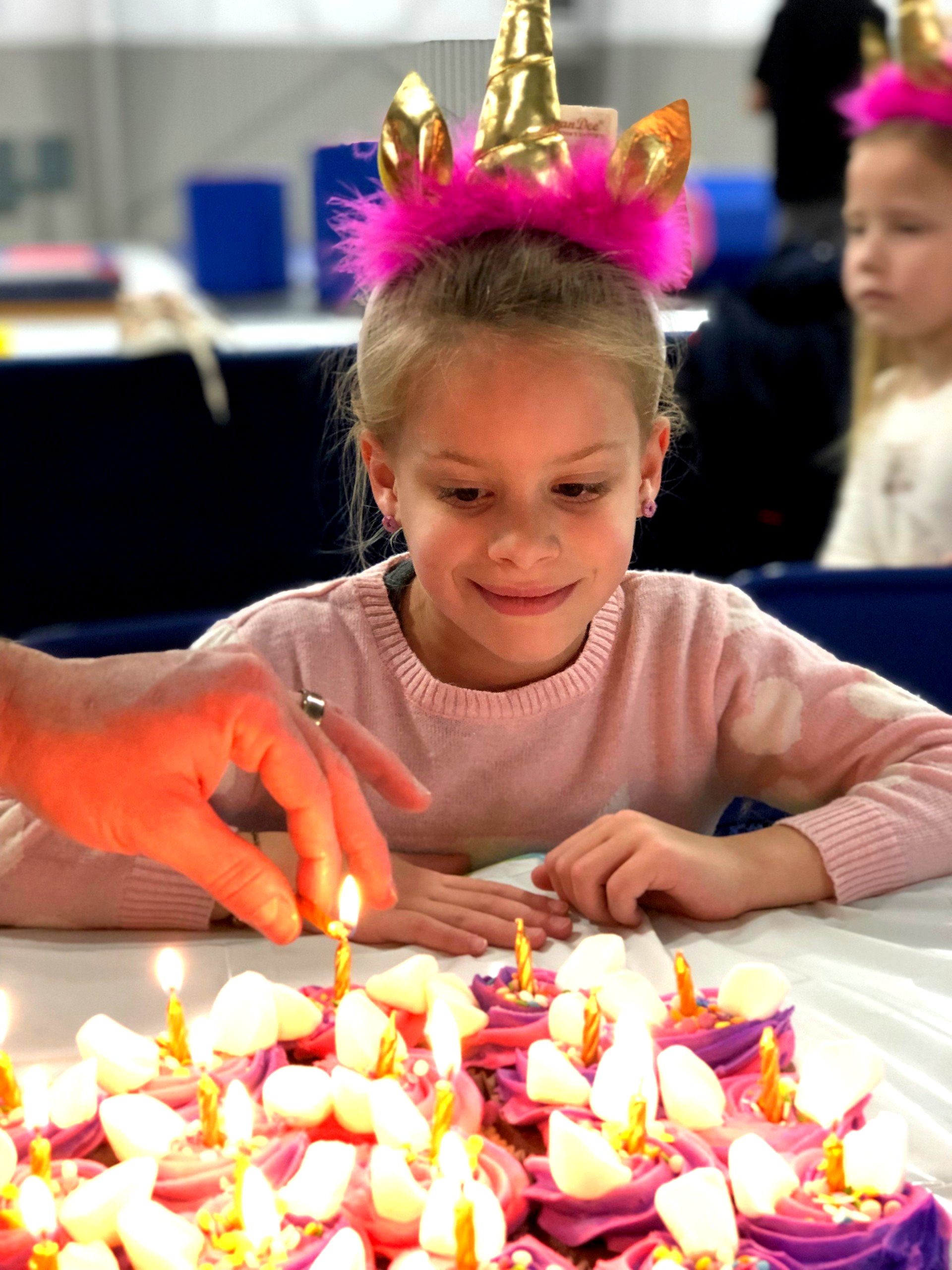 A little girl wearing a unicorn headband is sitting at a table with cupcakes and candles.