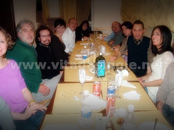 Un gruppo di persone è seduto attorno ad un lungo tavolo con sopra una bottiglia di vino