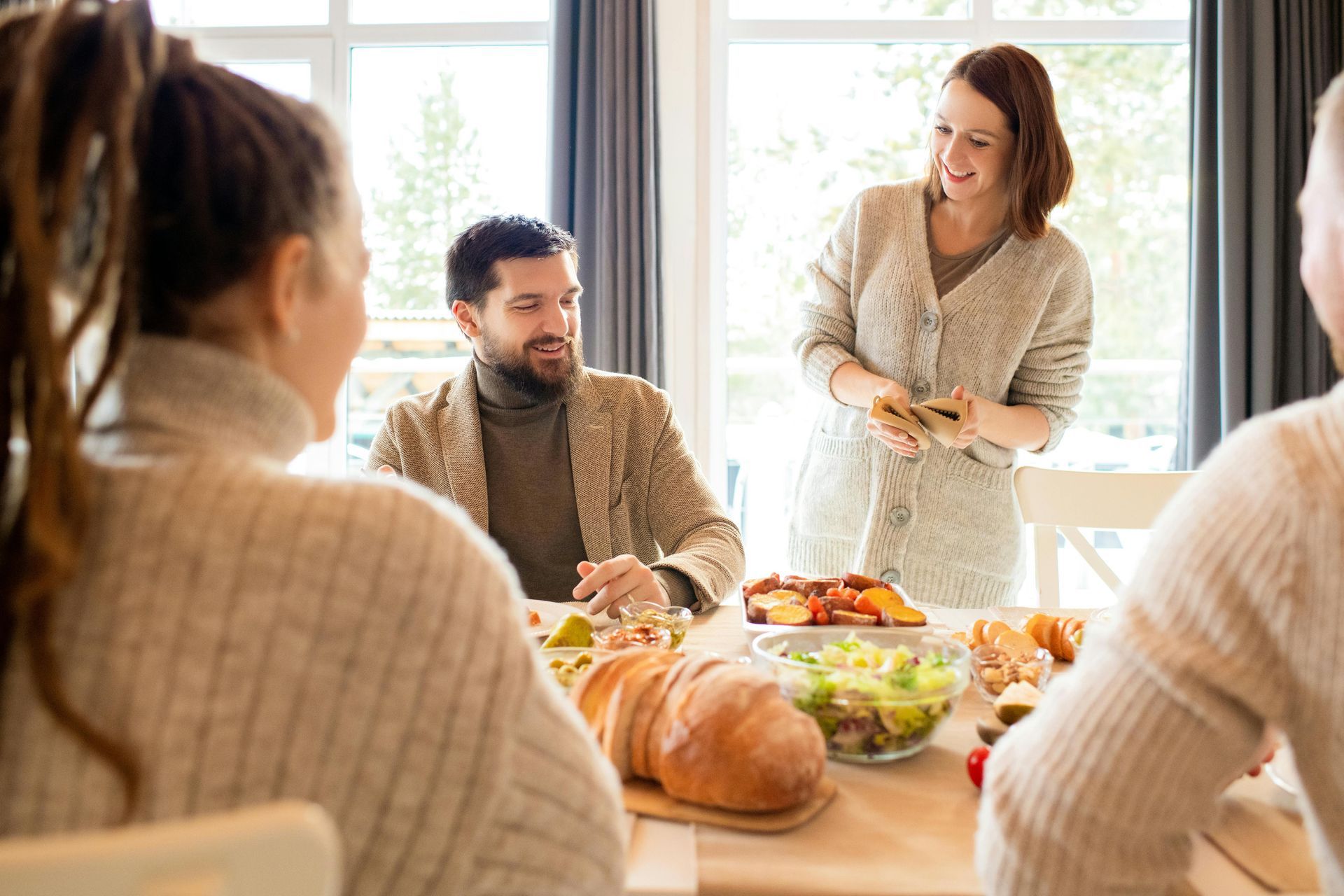 Persone attorno a un tavolo con del cibo, sorridenti. Una donna serve il cibo. Luce naturale e intensa.