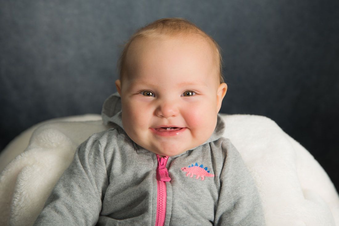 A baby is sitting on a white pillow and smiling at the camera.