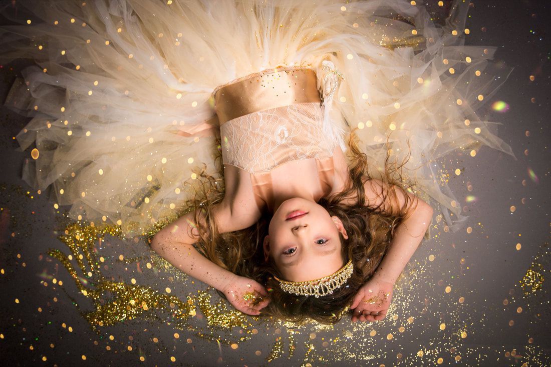 A little girl in a gold dress and tiara is laying on the floor surrounded by gold confetti.