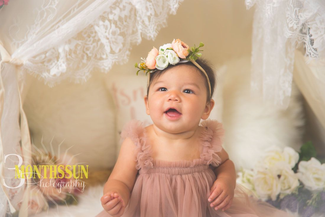 A baby girl wearing a pink dress and a flower crown is sitting in a teepee.