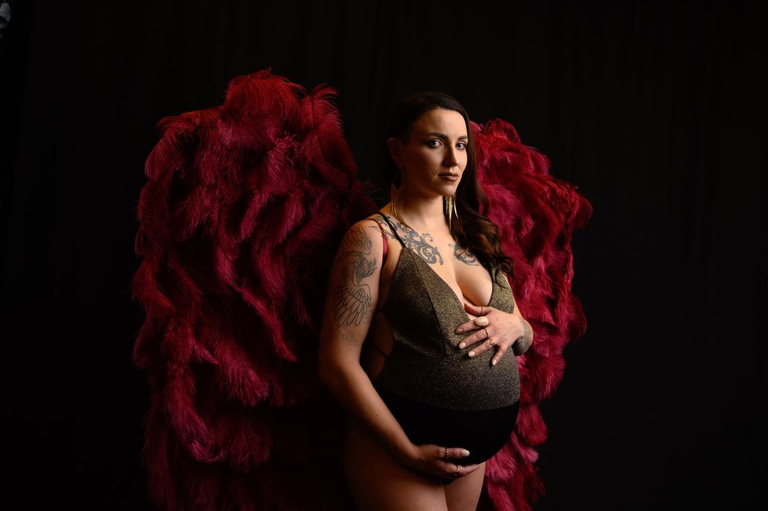 A pregnant woman is wearing red feather wings and holding her belly.