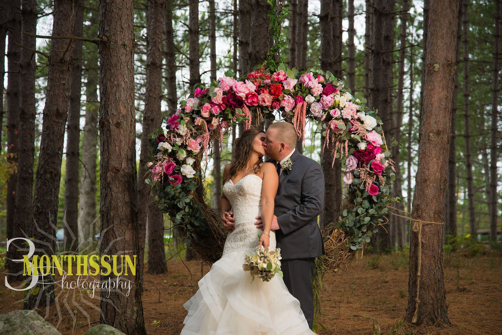 A bride and groom are kissing under a floral arch in the woods.