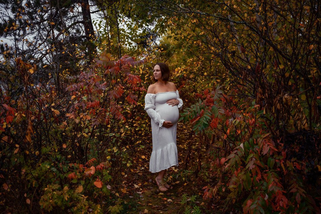 A pregnant woman in a white dress is standing in the woods holding her belly.