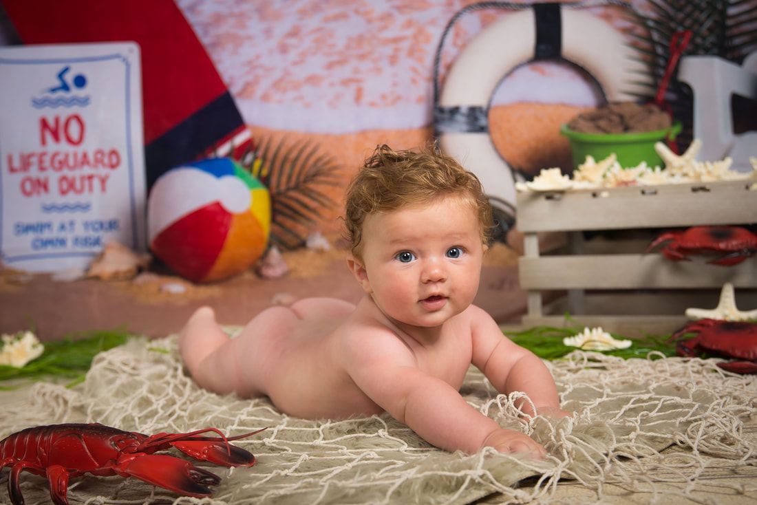 A baby is laying on a beach with a lifeguard on duty sign in the background.