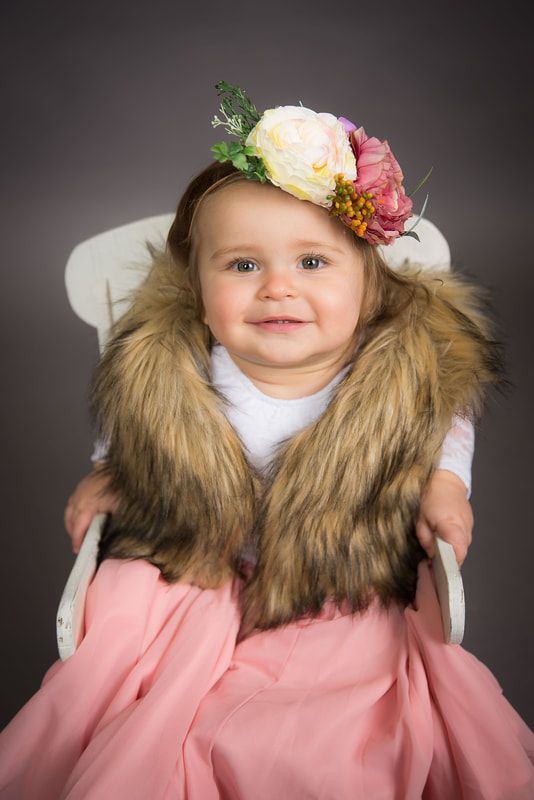 A little girl is sitting in a chair wearing a fur vest and a flower crown.