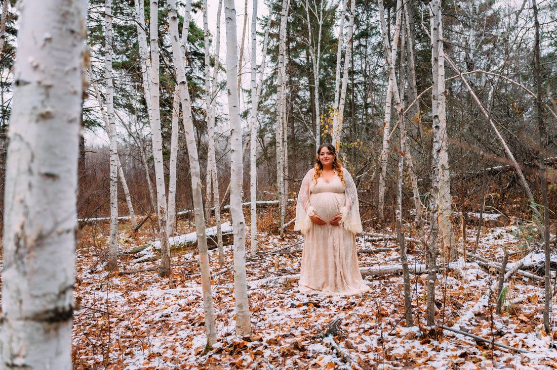 A pregnant woman in a white dress is standing in a snowy forest.
