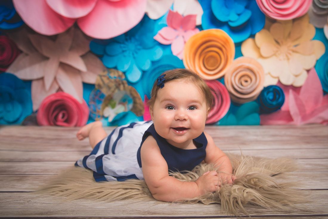 A baby girl is laying on a wooden floor in front of a wall of paper flowers.
