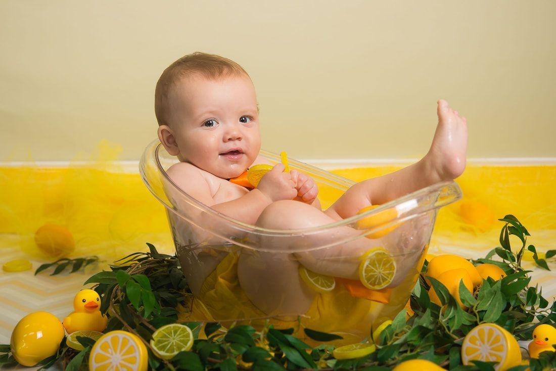 A baby is taking a bath in a bowl filled with lemons and rubber ducks.
