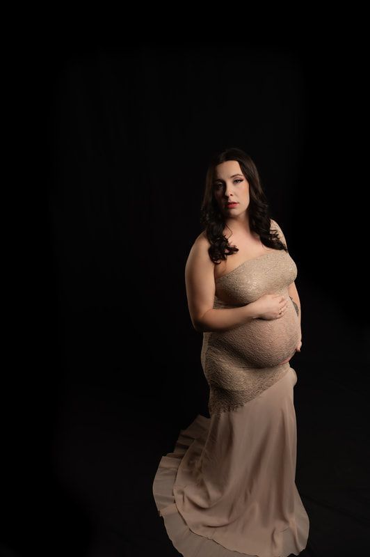 A pregnant woman in a long dress is standing in front of a black background.