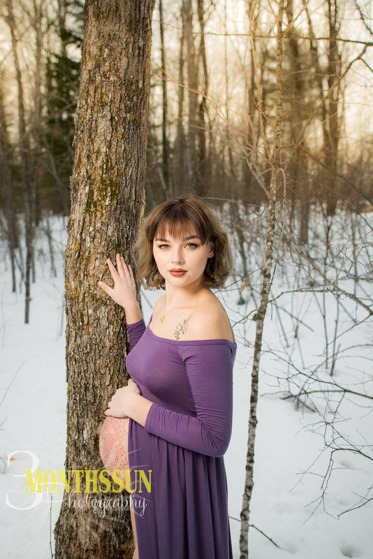 A pregnant woman in a purple dress is standing next to a tree in the snow.