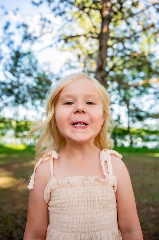 A little girl in a white dress is standing in front of a tree in a park.