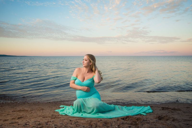 A pregnant woman in a blue dress is kneeling on the beach.