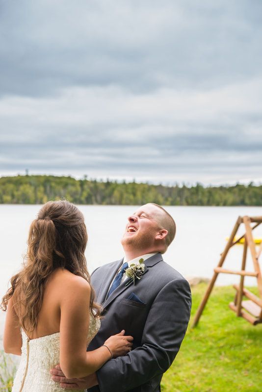 A bride and groom are laughing in front of a lake.