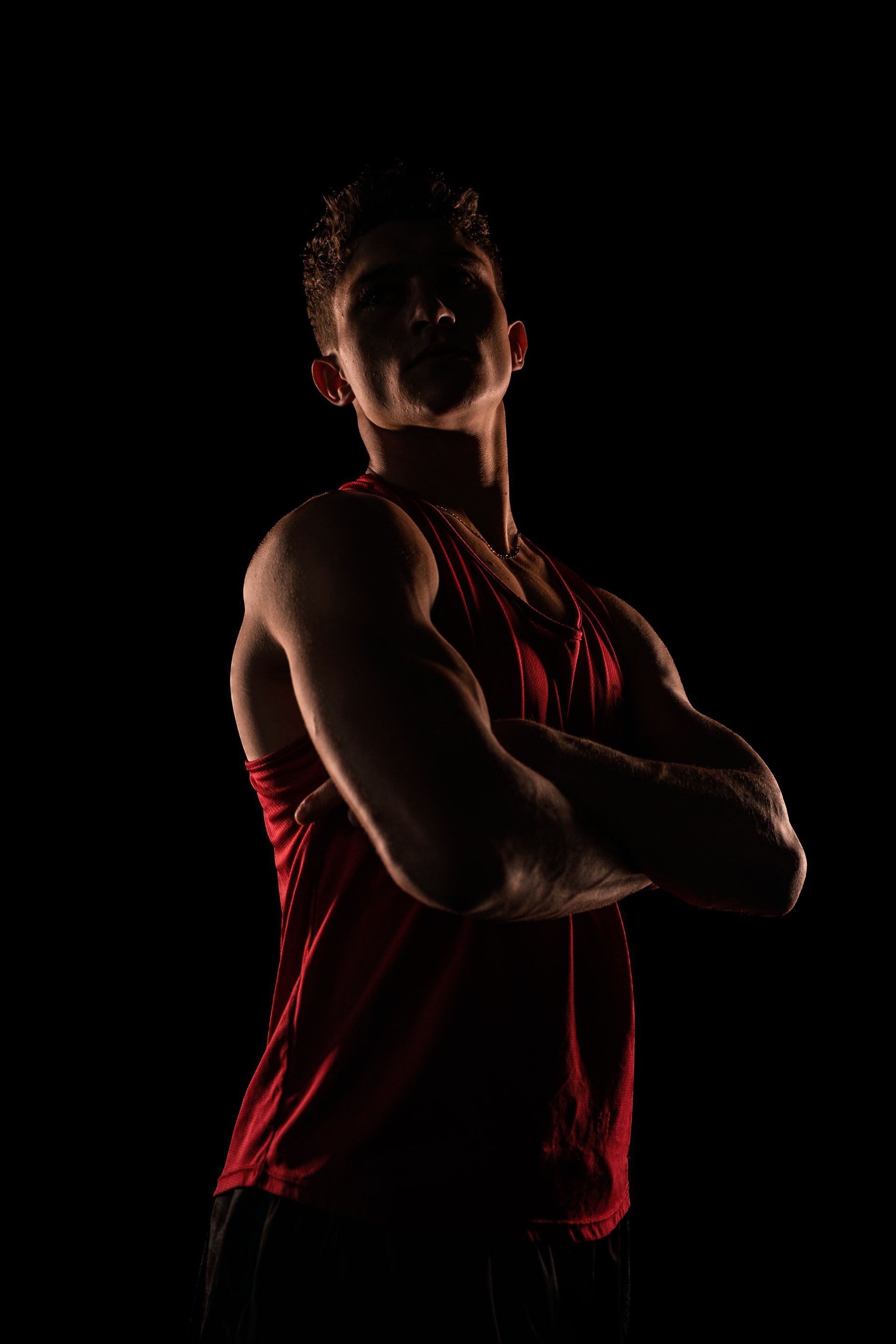 A man in a red tank top is standing in the dark with his arms crossed.