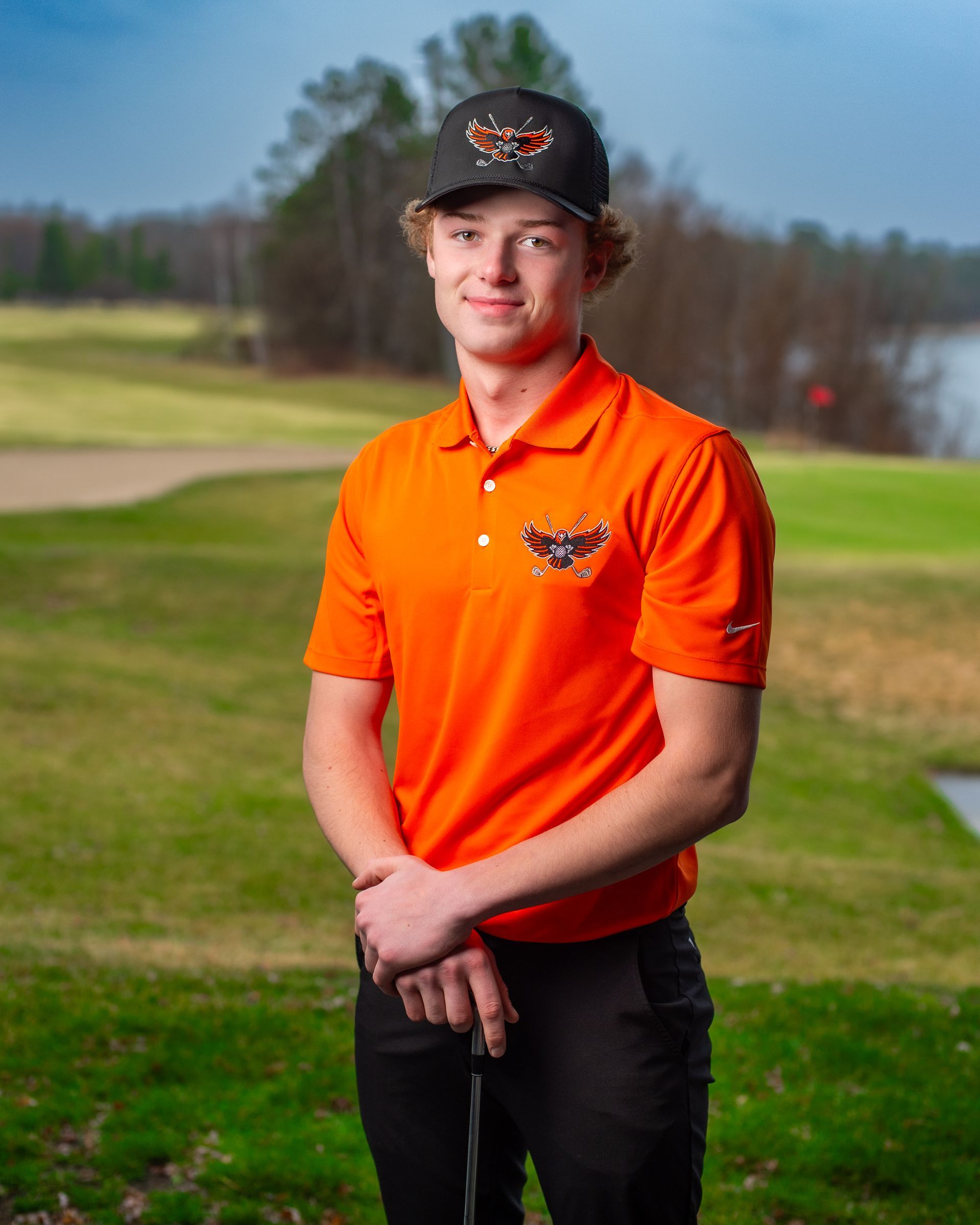 A young man in an orange shirt and black hat is standing on a golf course holding a golf club.