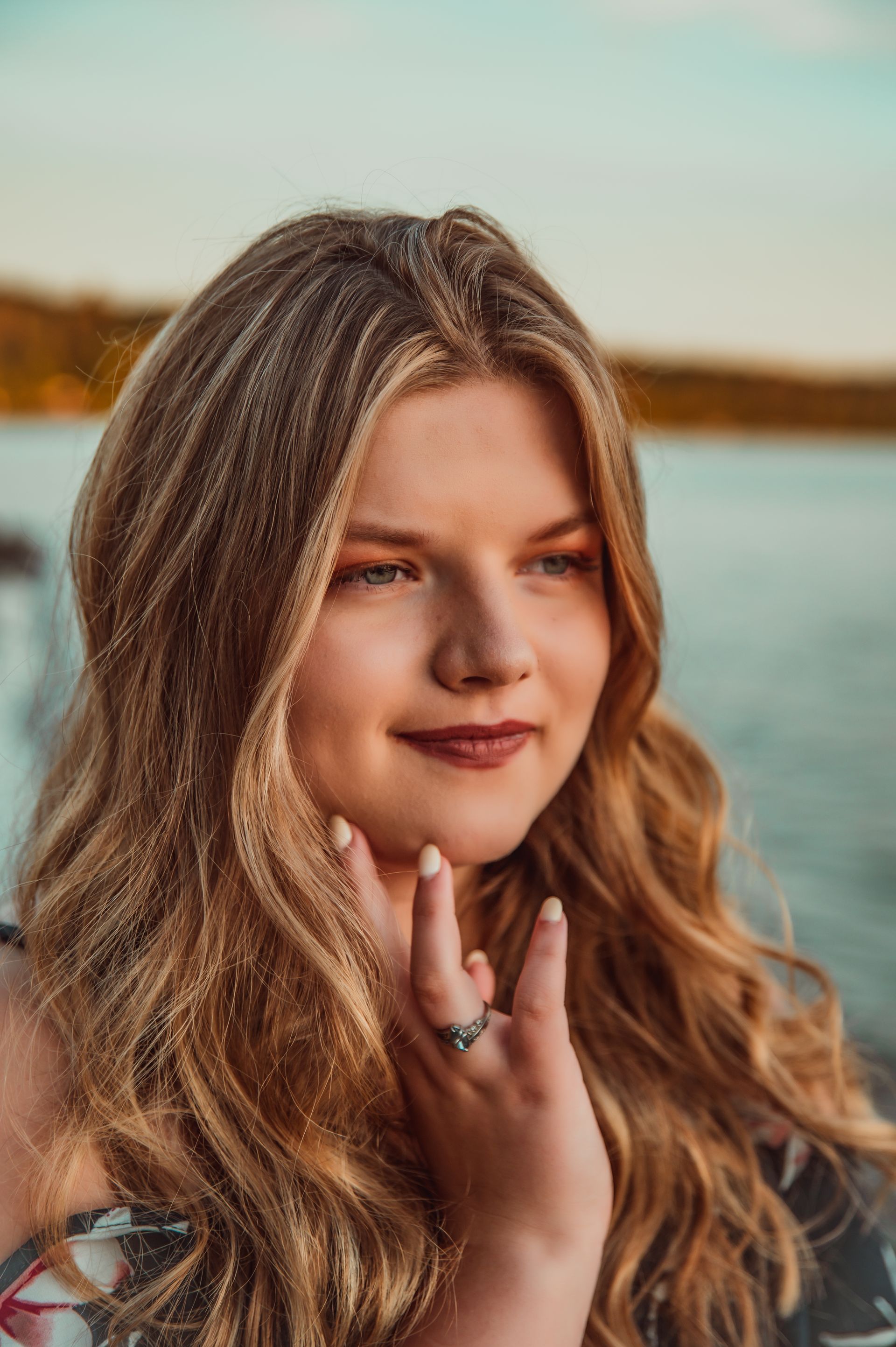 A woman with long hair and a ring on her finger is standing in front of a body of water.