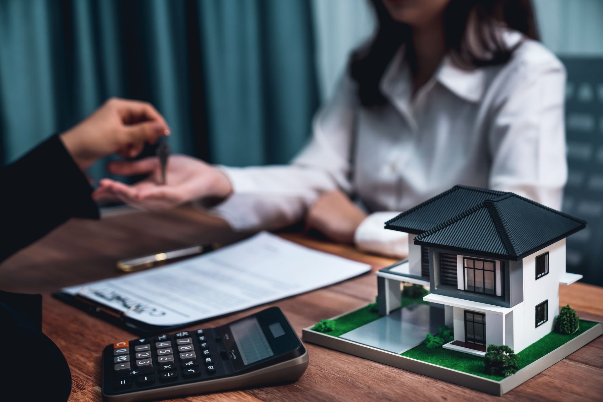 A woman is sitting at a table with a model house and a calculator.