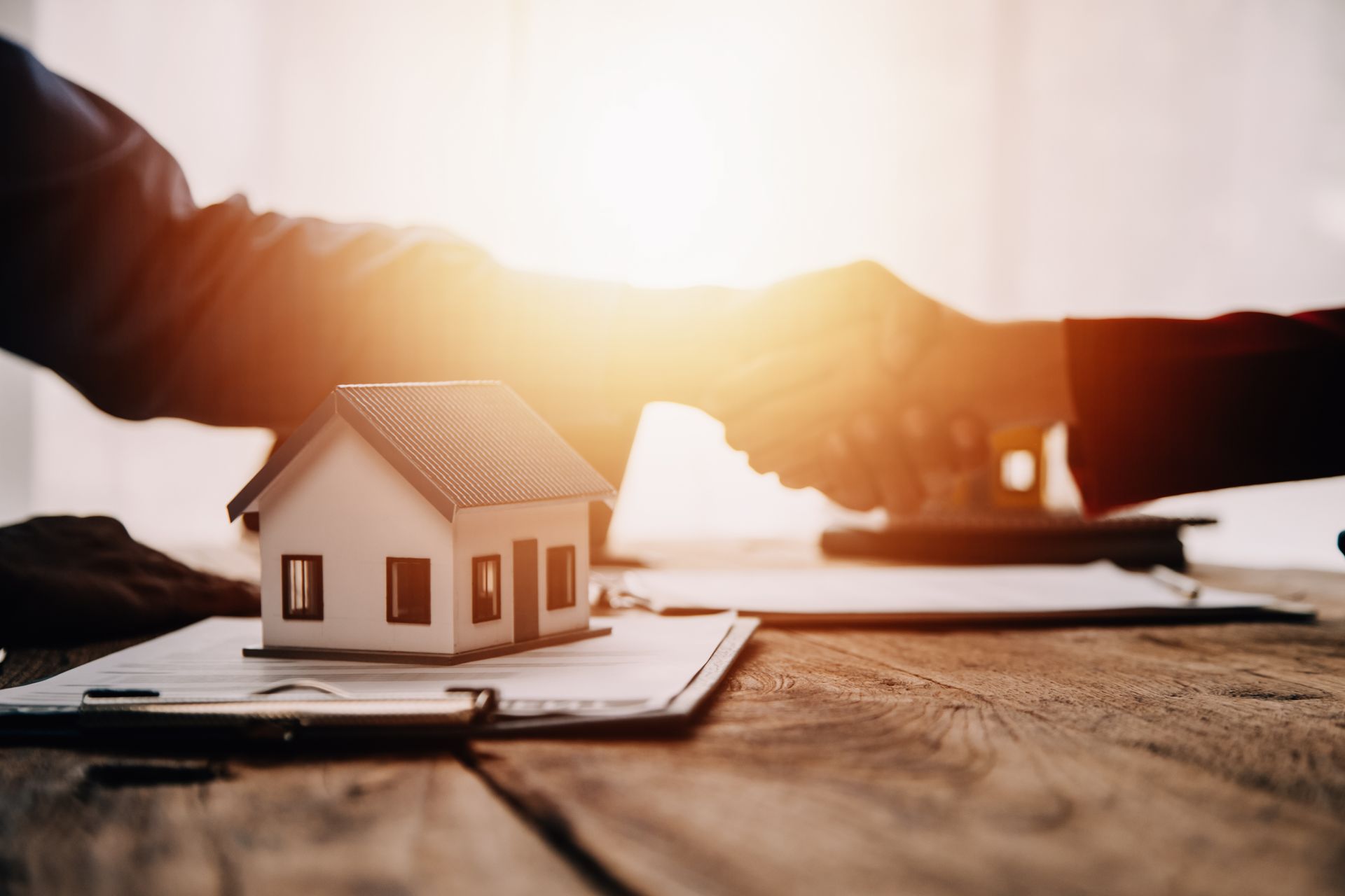 Two people are shaking hands over a model house on a clipboard.