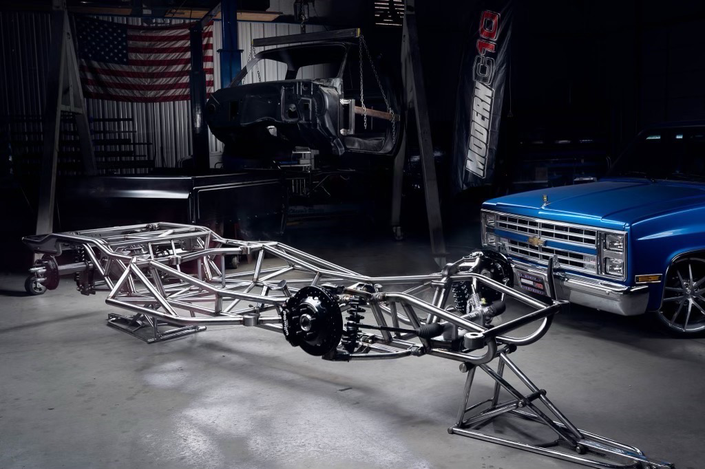 Steel car chassis on a stand, with a blue truck and a car body in a garage. An American flag hangs on the wall.