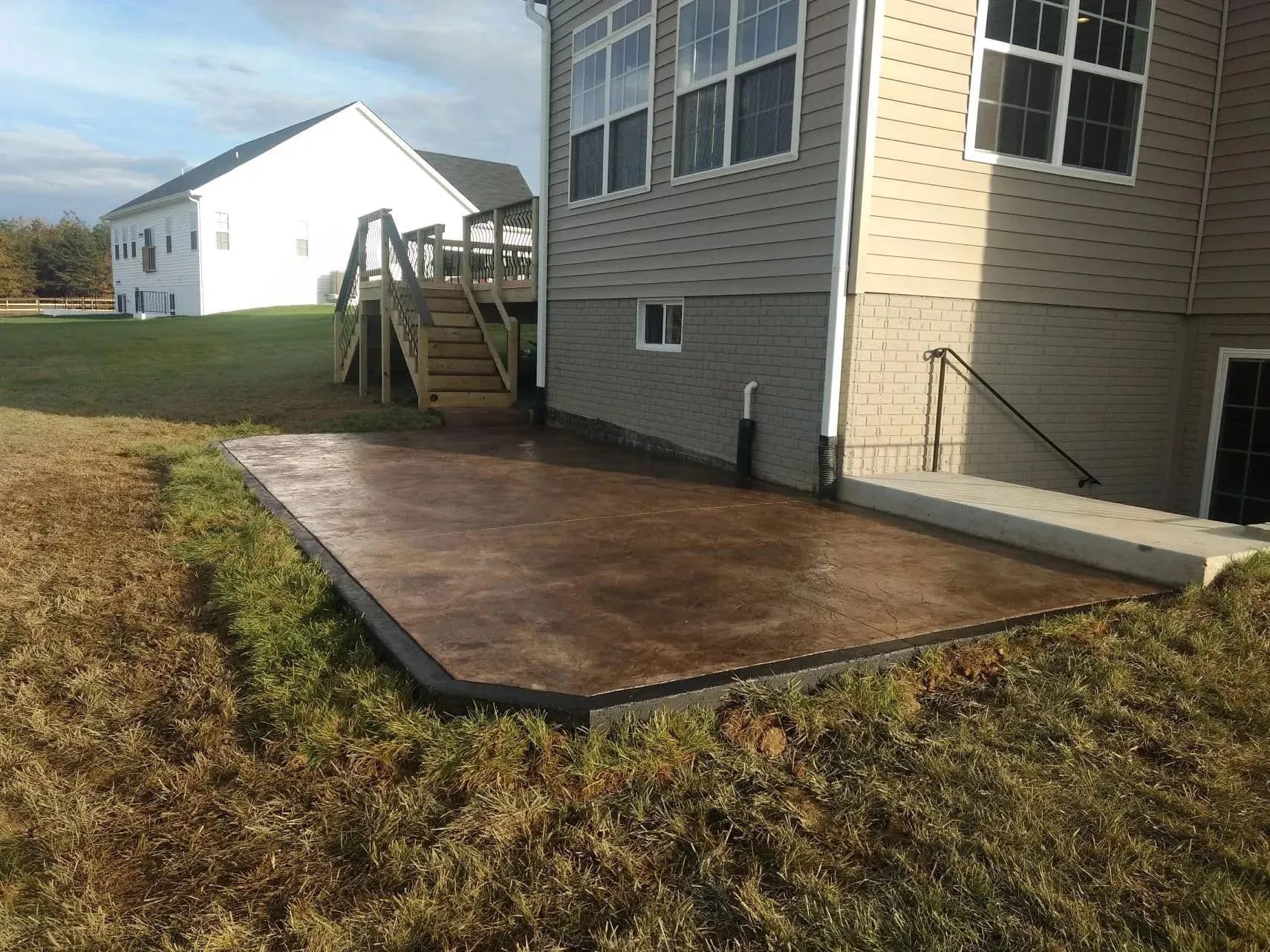 Brown concrete patio next to a house with stairs. Grassy yard.