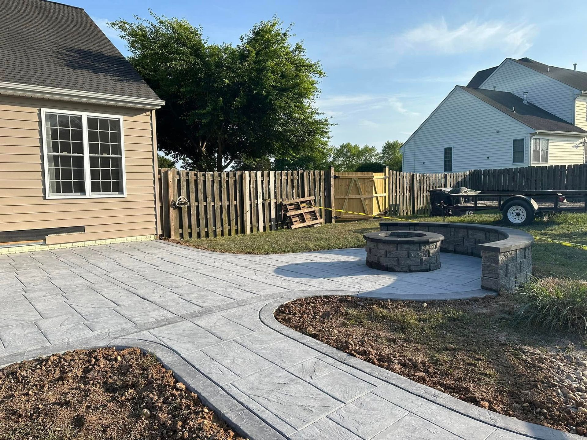A patio with gray pavers and a stone fire pit in a residential backyard with a house, wooden fence, and nearby trailer.