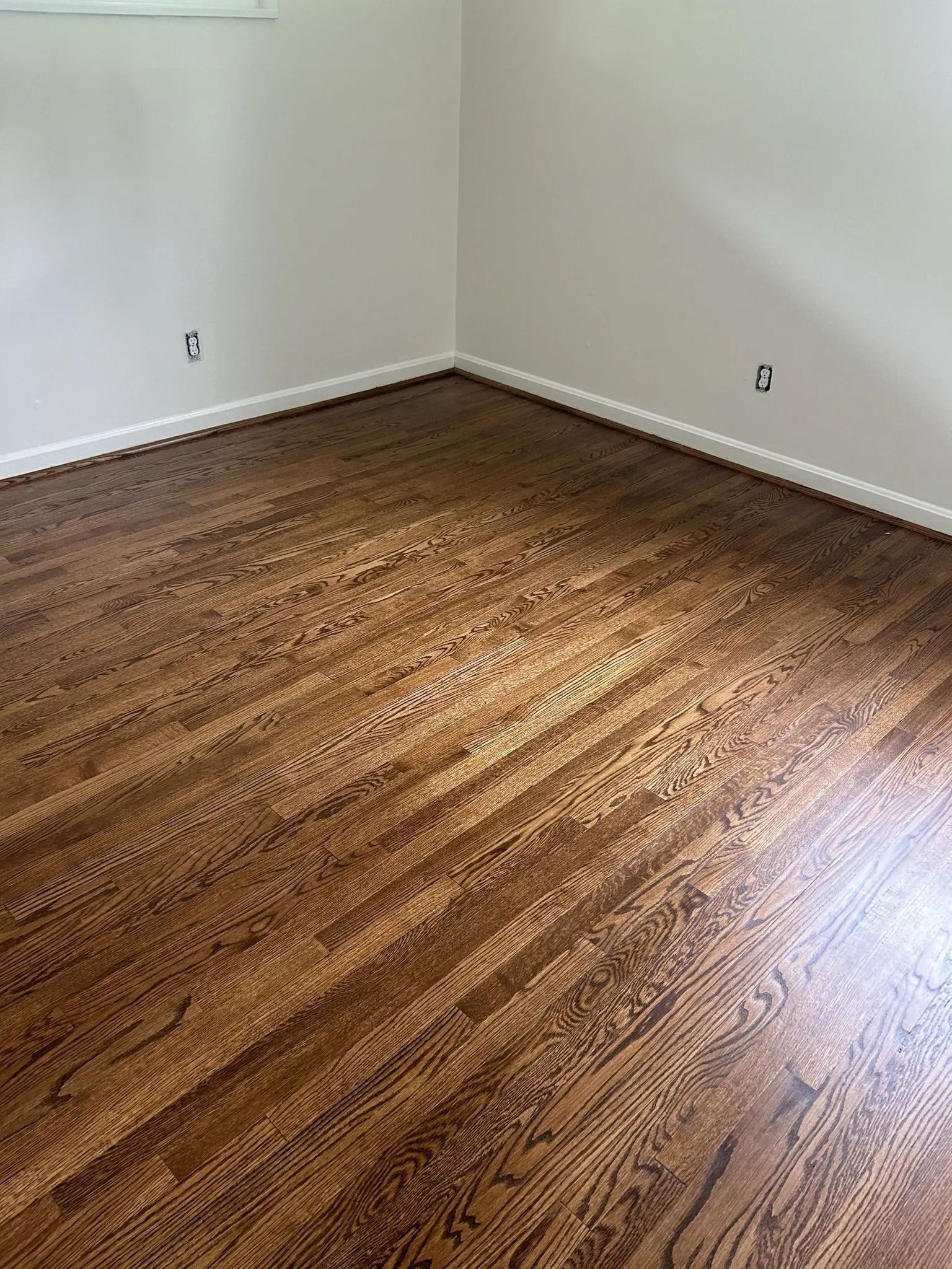 Dark stained hardwood floor in a corner of a room with white walls and baseboards.