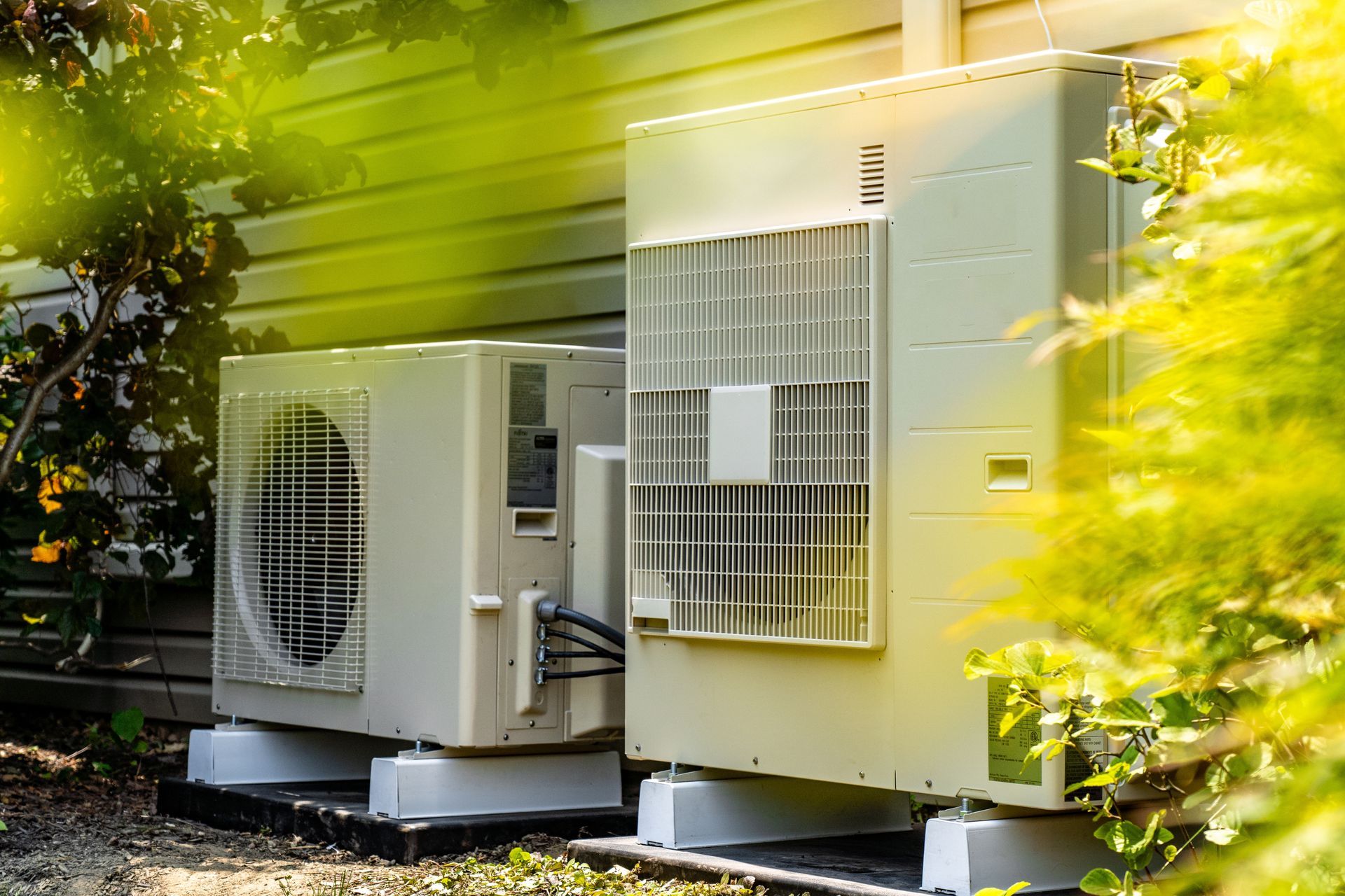 Two Beige Air Conditioning Units Sit Outside, Partially Obscured by Green Foliage — Carl Voss-Electrician In Urangan, QLD