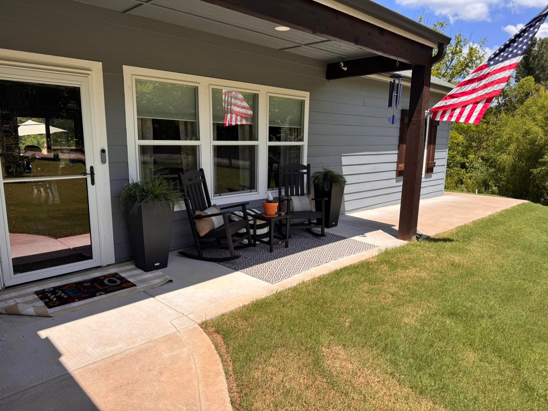 A porch with a grey house, black rocking chairs, an American flag, and green grass.