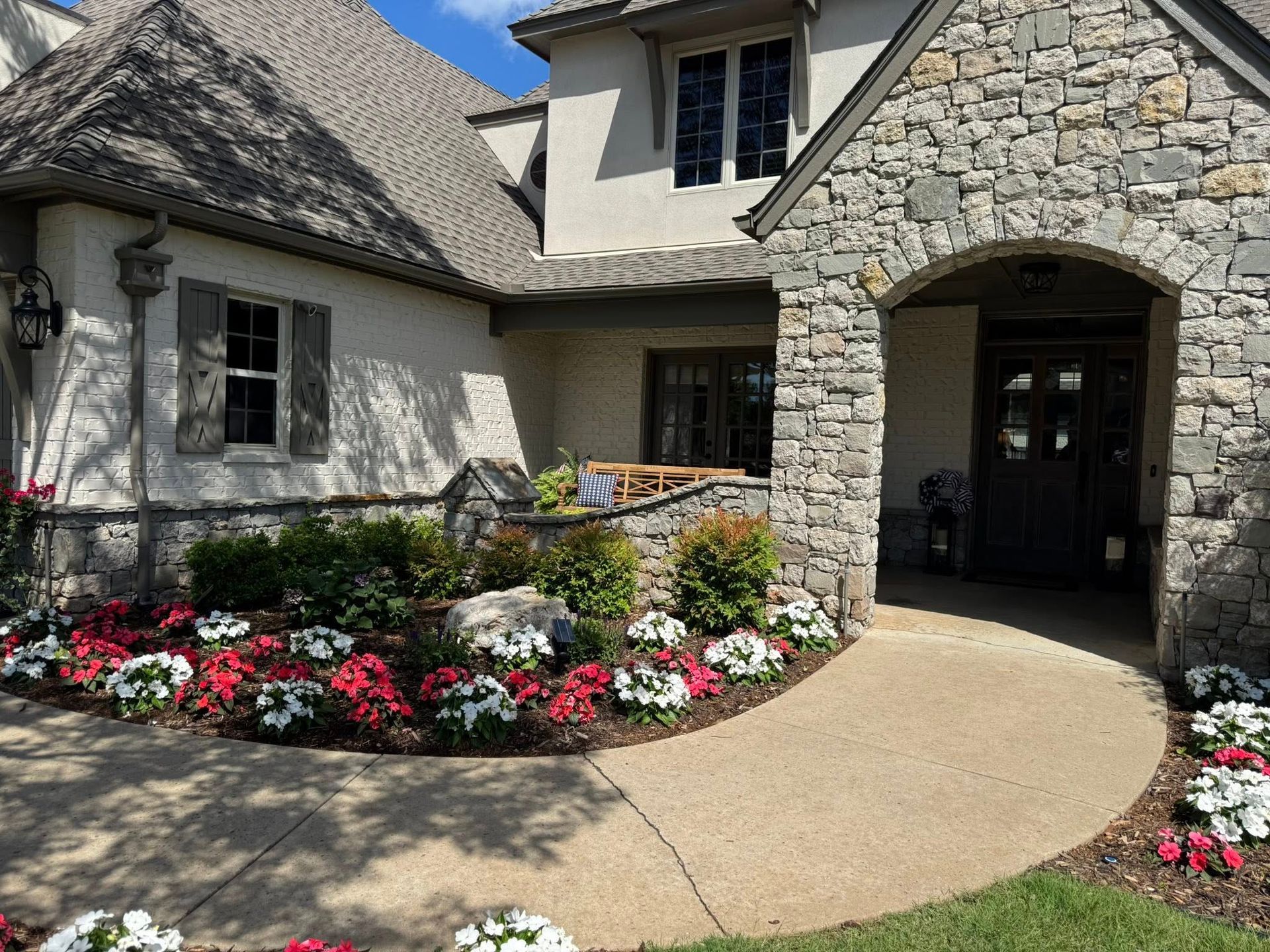 Stone-faced house with arched entry and colorful flower beds along a curved walkway.