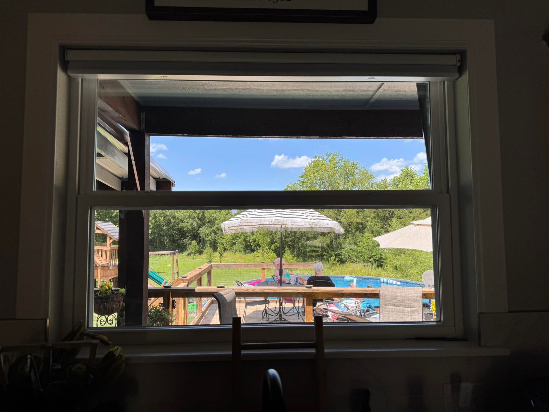 View through a window: blue sky, backyard with pool, trees, and outdoor furniture.