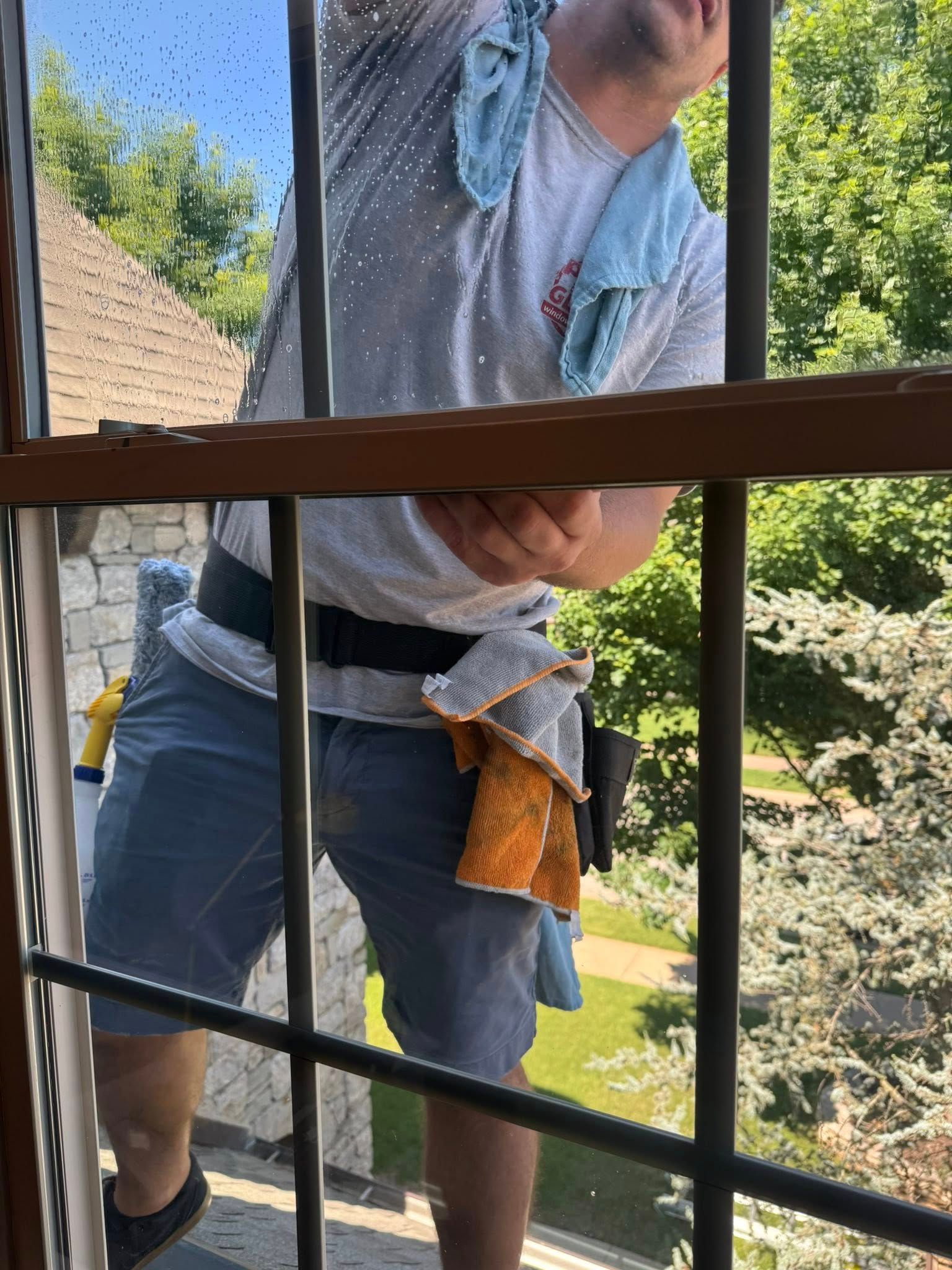 Man washing window, wearing gray shirt and blue shorts, outside near trees.