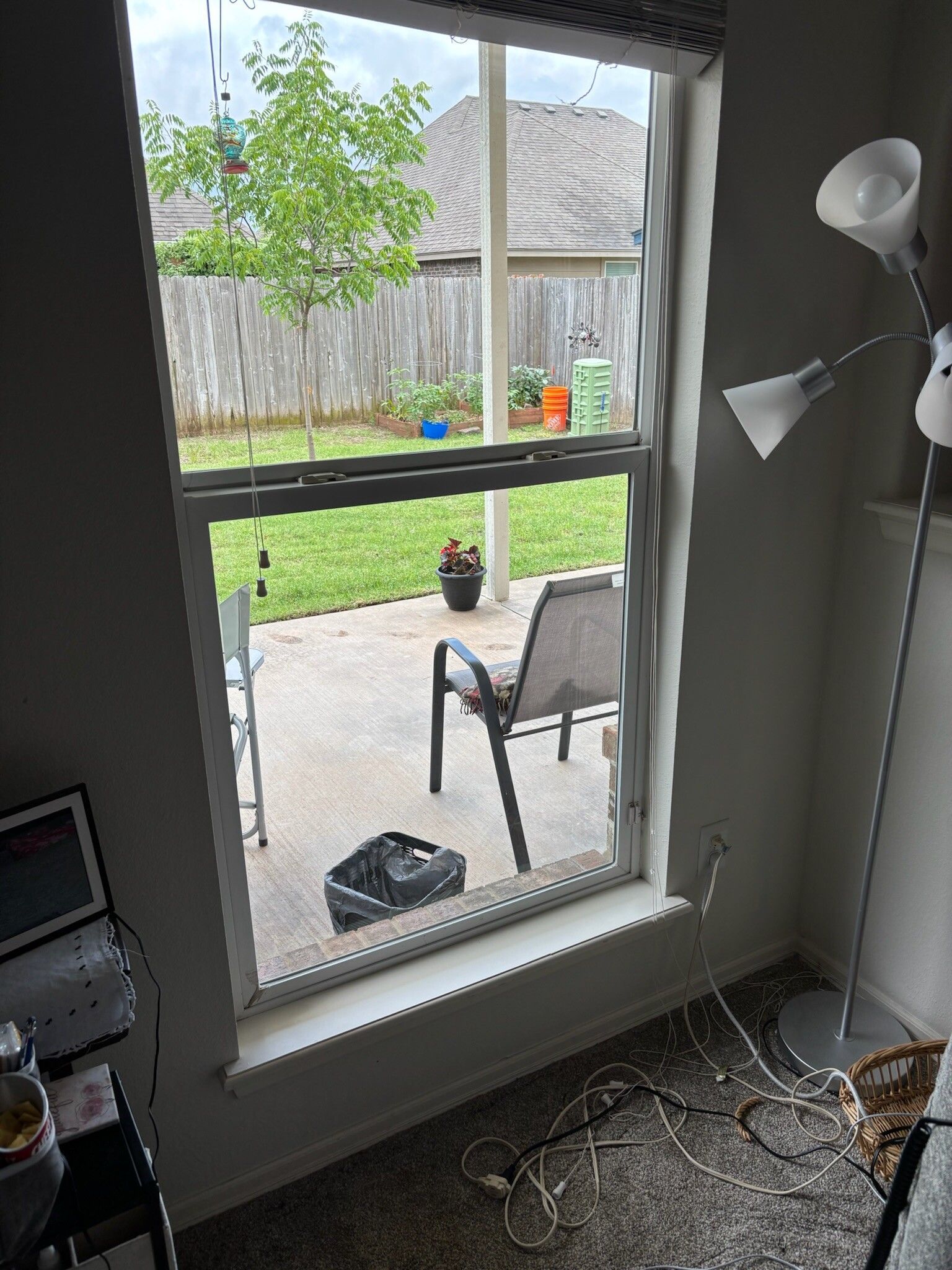Window view of a patio with a chair, a tree, and a green yard. A lamp stands beside the window.